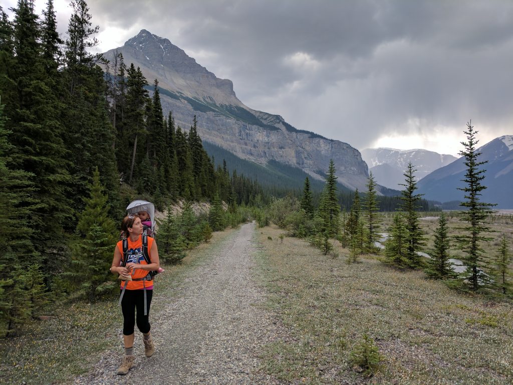 A hiker with a small child on her back walking down a path heading away from mountains