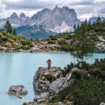 A hiker descending a large rock at the edge of Lago di Sorapis with mountains in the background