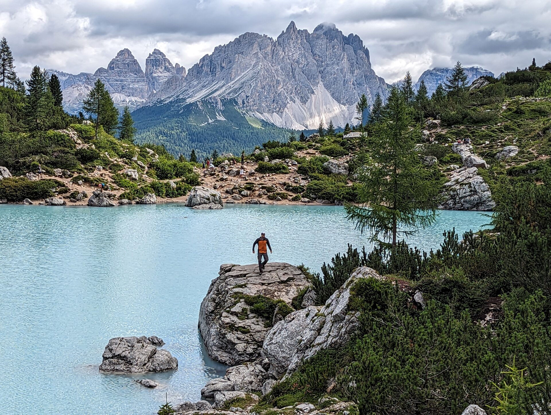A hiker descending a large rock at the edge of Lago di Sorapis with mountains in the background