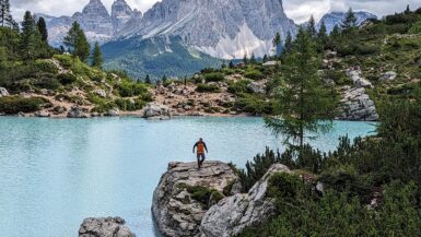 A hiker descending a large rock at the edge of Lago di Sorapis with mountains in the background