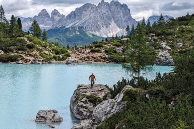 A hiker descending a large rock at the edge of Lago di Sorapis with mountains in the background