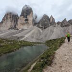 A hiker on a path beside water and the cloud-covered Tre Cime formation