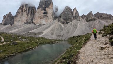 A hiker on a path beside water and the cloud-covered Tre Cime formation