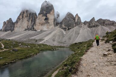 A hiker on a path beside water and the cloud-covered Tre Cime formation