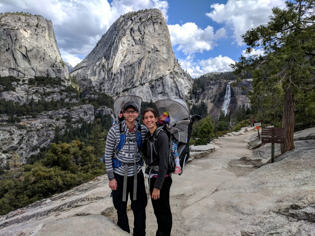 A hiking couple carrying kids in backpacks with a waterfall and large rock formation in the background