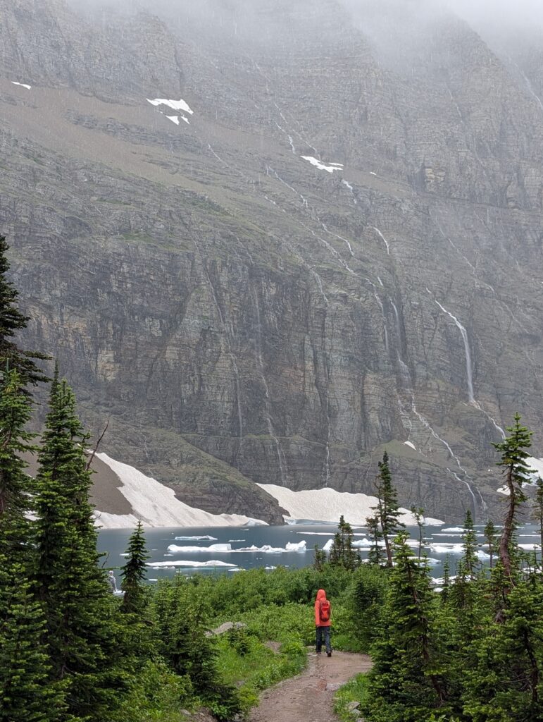 A hiker following a curved path with a pool of water and looming, cloud-covered mountains in the background