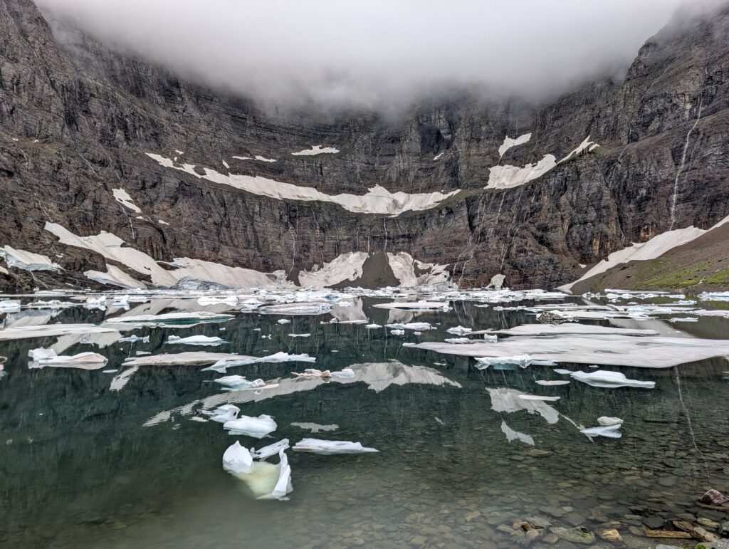 Several thin waterfalls running down a mountain wall into a pool of water that has many icebergs at Iceberg Lake
