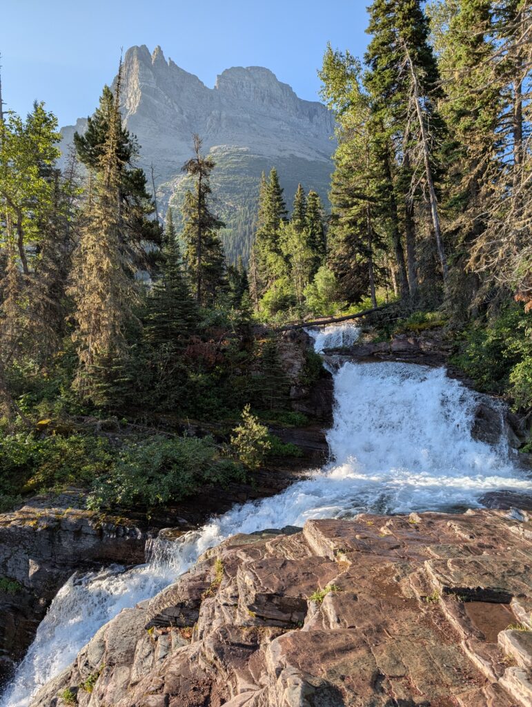 A waterfall surrounded by pine trees flows down and across a rocky mountain landscape