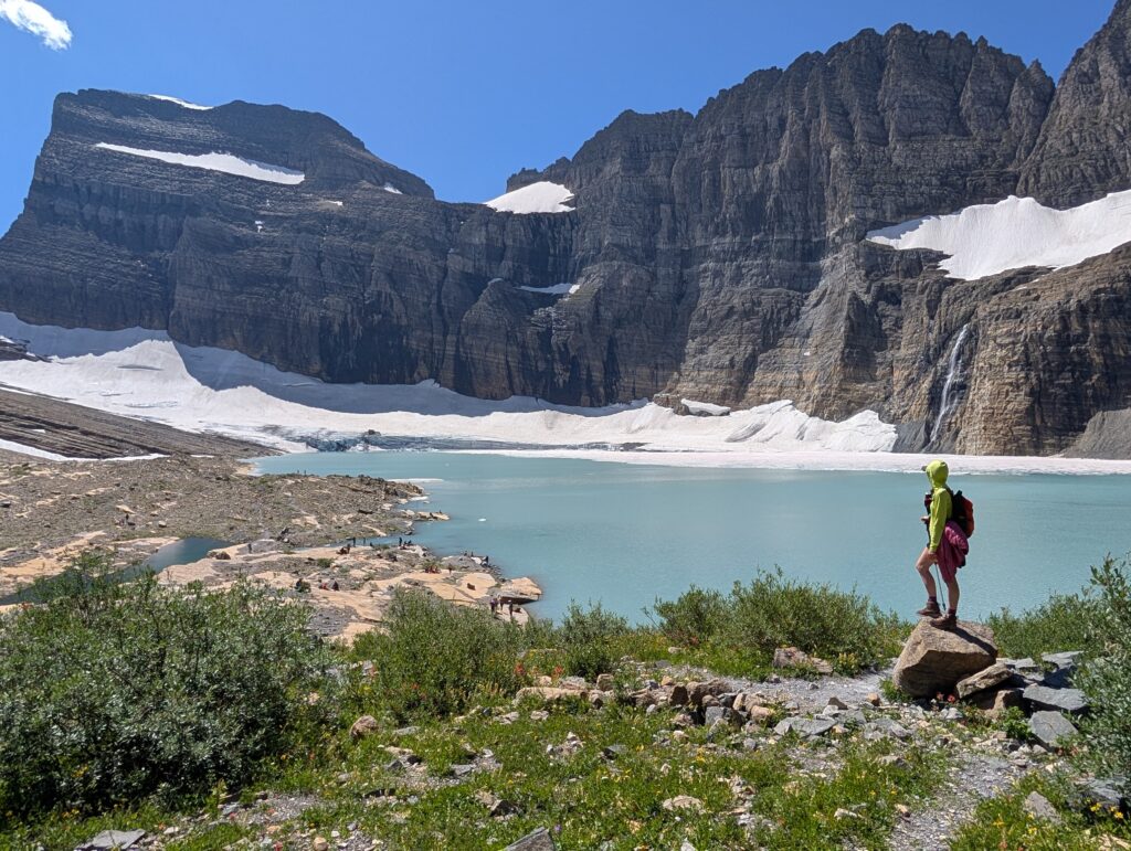 A hiker stands on a rock overlooking a glacier feeding a pool of water in Glacier National Park