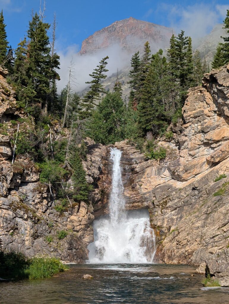 A narrow waterfall meeting a thicker waterfall before plunging into a pool with cloudy mountains above it