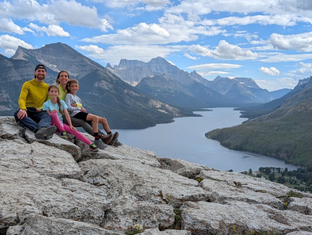 A family of 4 sitting on a rock with a body of water lined with mountains on both sides