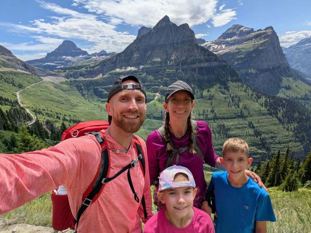 A family of four with multiple mountain peaks behind them on the Highline Trail