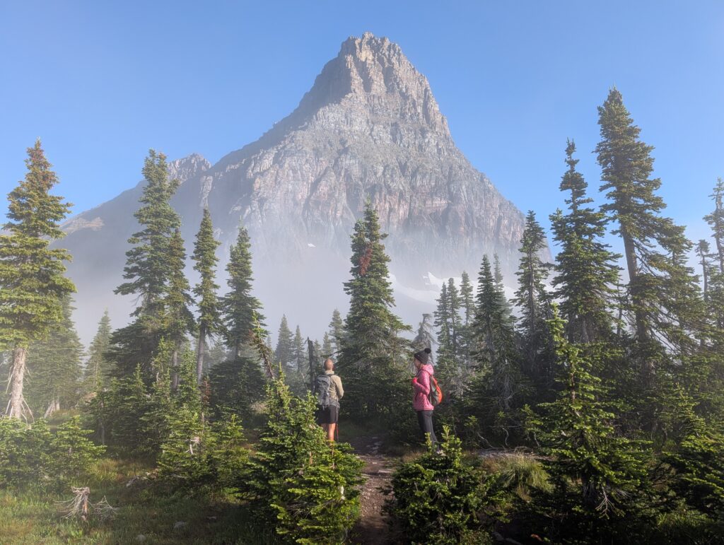 Two hikers standing among pine trees with a cloudy tall mountain peak in the background