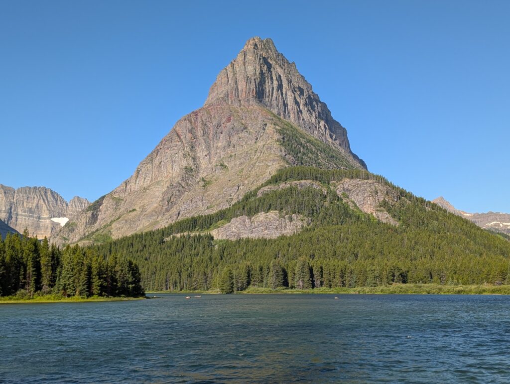 A pointy mountain in front of a body of water in Many Glacier