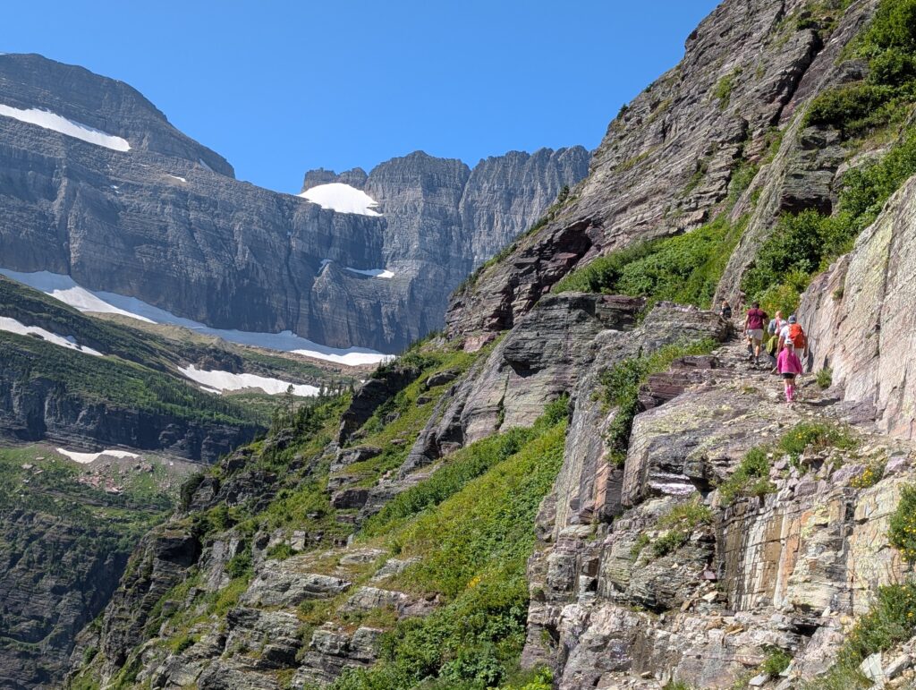 Hikers along a narrow stone path with patches of snow in the mountains in the background