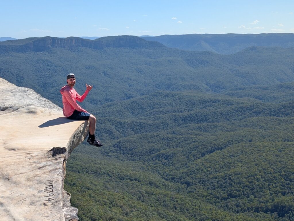 A man sitting at the very edge of a high cliff with a green rolling mountains in the background