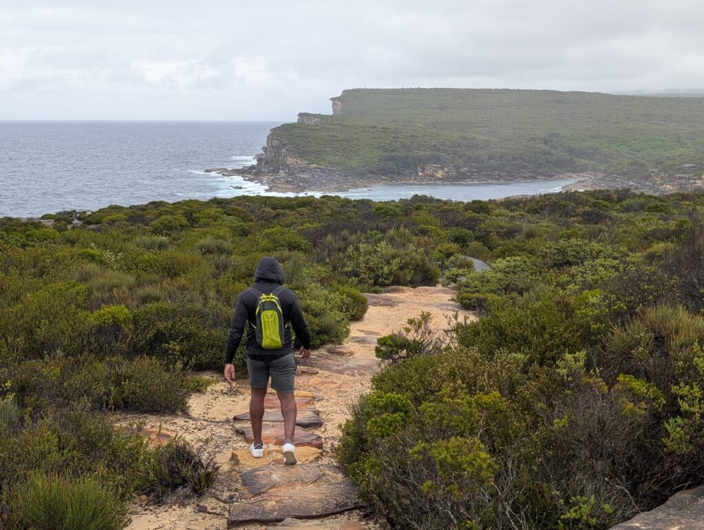 The back of a man walking along a sandy, rocky trail with a jagged coastline in the distance
