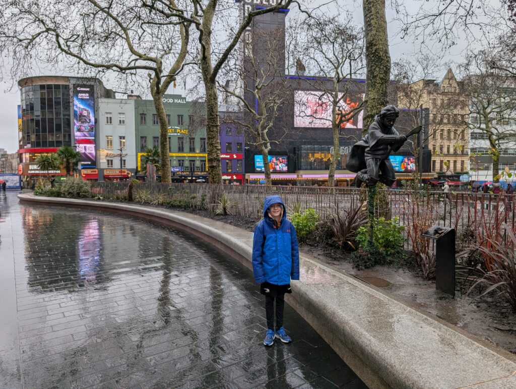 A young boy standing by the Harry Potter statue in Leicester Square, London on a rainy day
