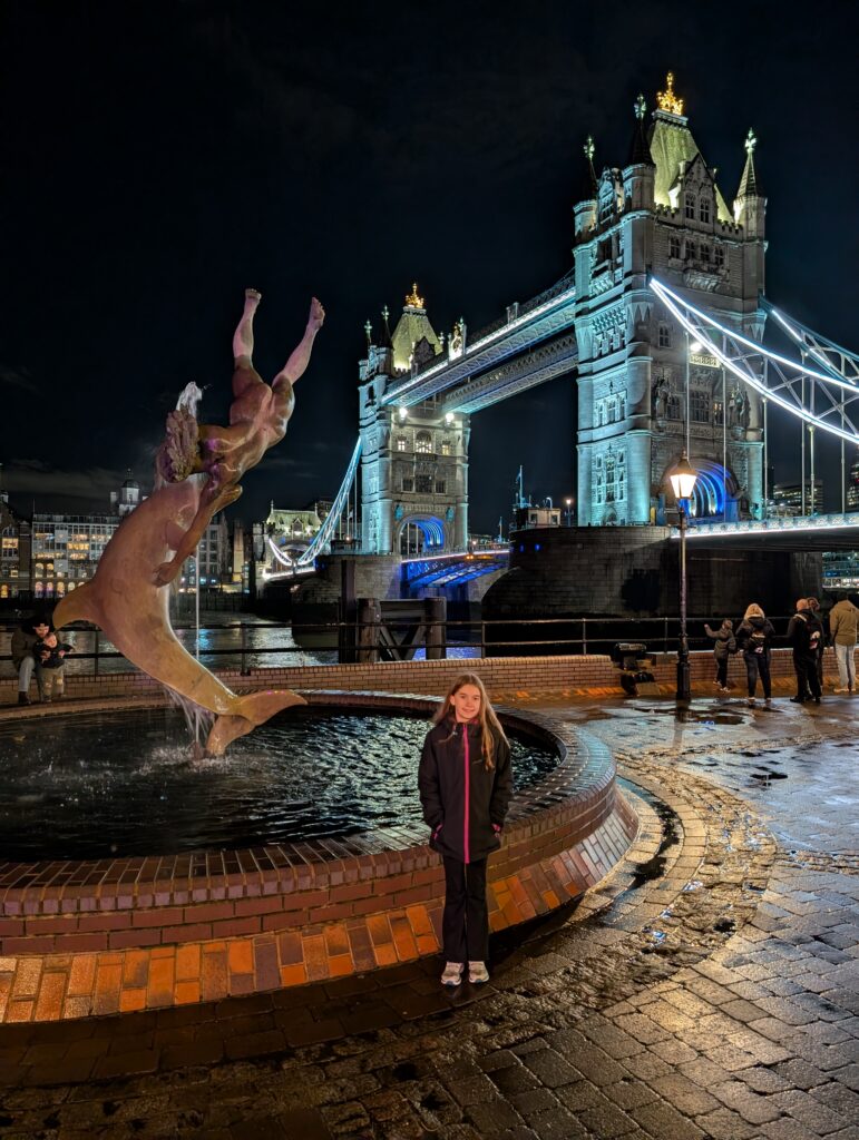 A young girl in front of a fountain with a dolphin sculpture and large lit-up bridge in the background