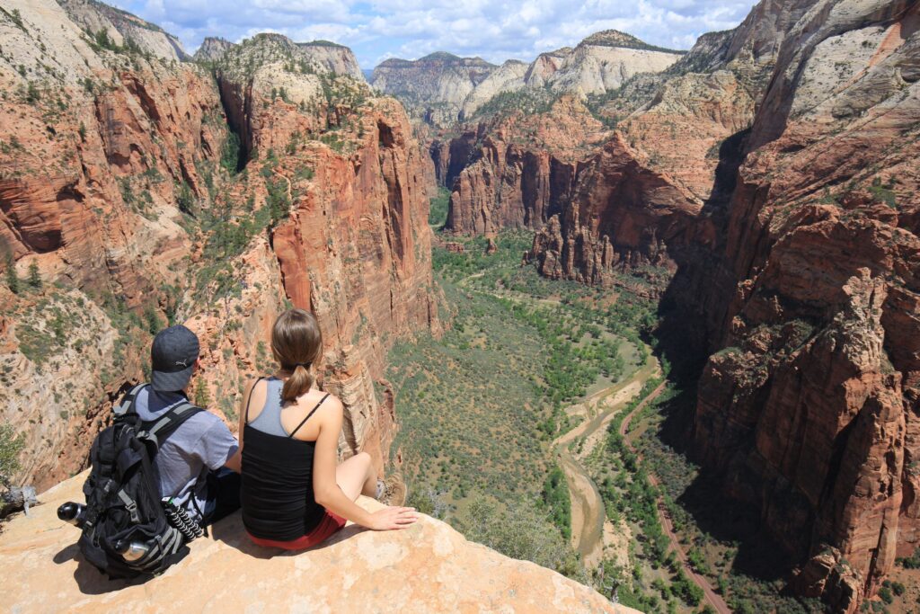 A man and woman sit on a rock ledge looking out at a canyon far below