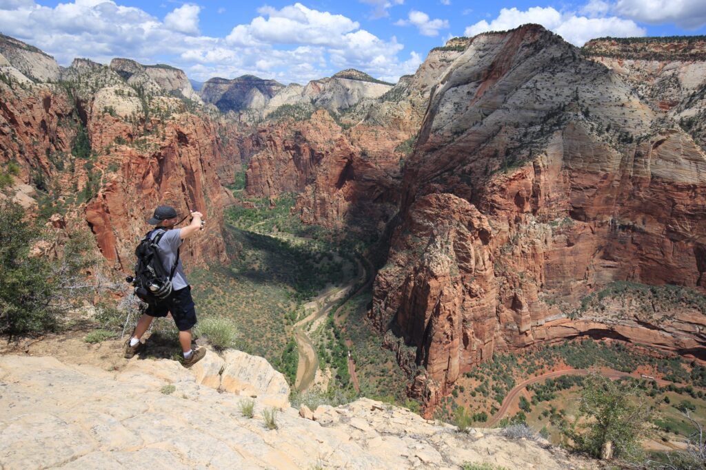 A man taking a picture on a ledge with a green and red canyon in the background