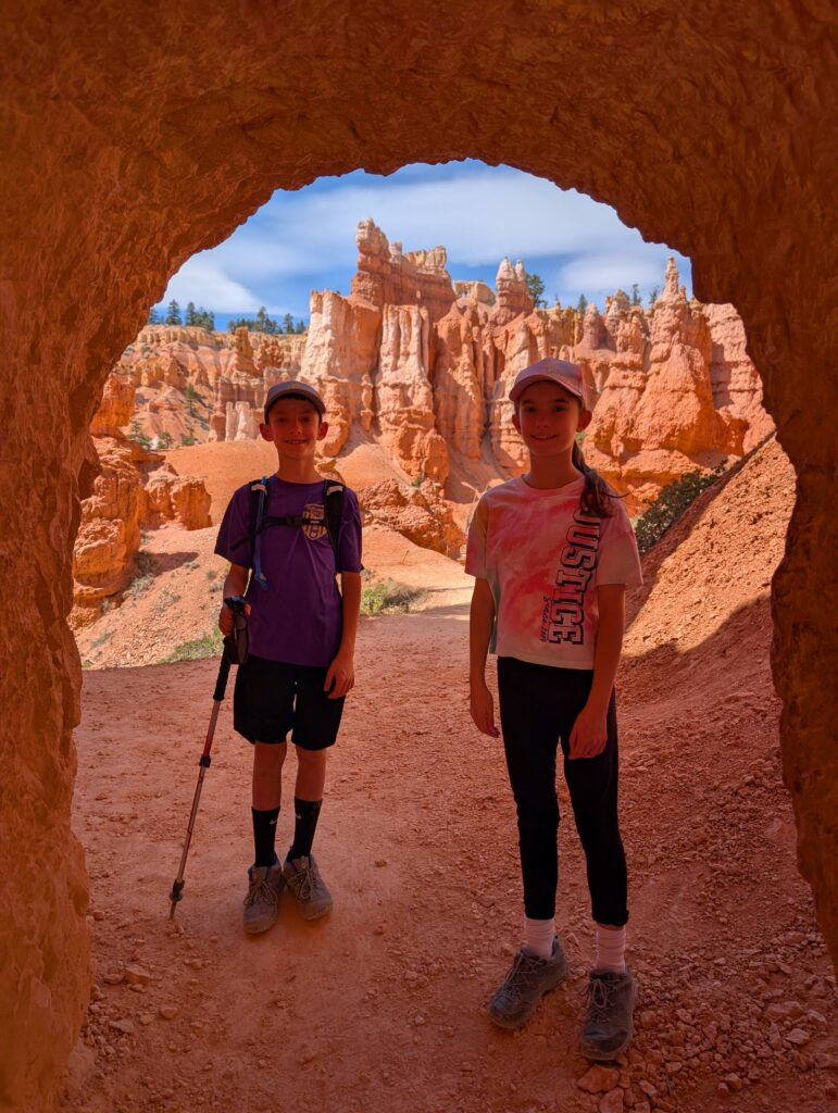A boy and girl standing at the end of a stone tunnel with rock formations framed behind them