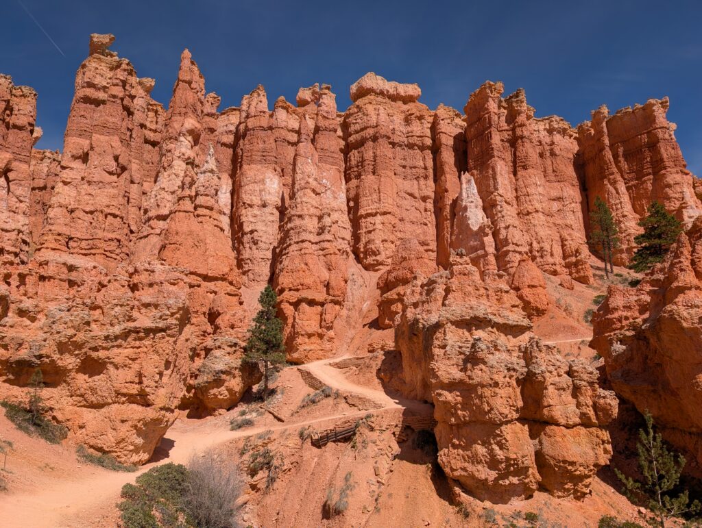 An orange rock wall with a sandy hiking path zig-zagging at the bottom of it