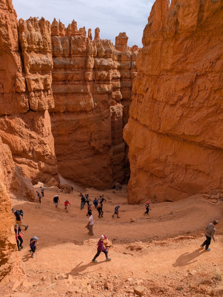 People climbing sandy switchbacks between red-orange cliff walls