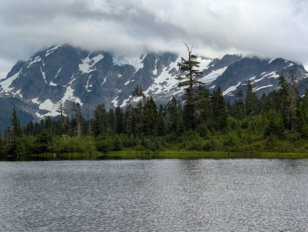 Clouds covering the top half of a mountain with trees and water in the foreground