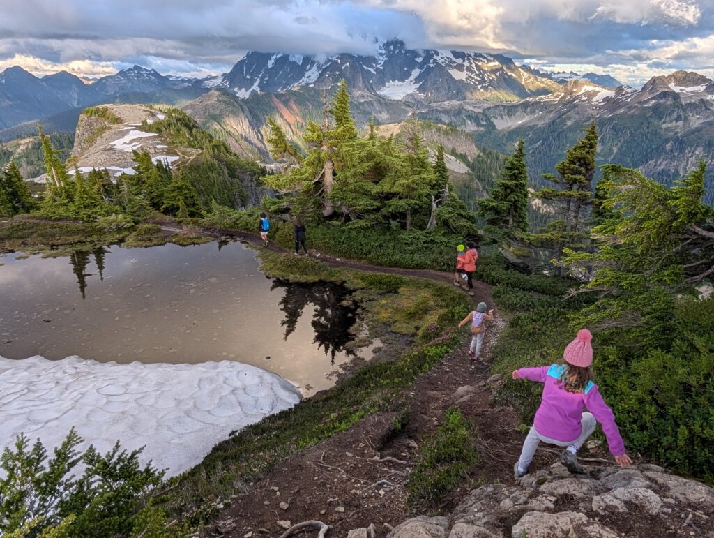 Hikers navigating a path that loops around a small snow-fed body of water with mountains and trees