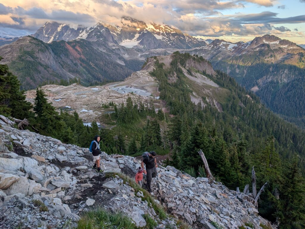 A father and 2 boys walking along loose rocks with mountains and clouds