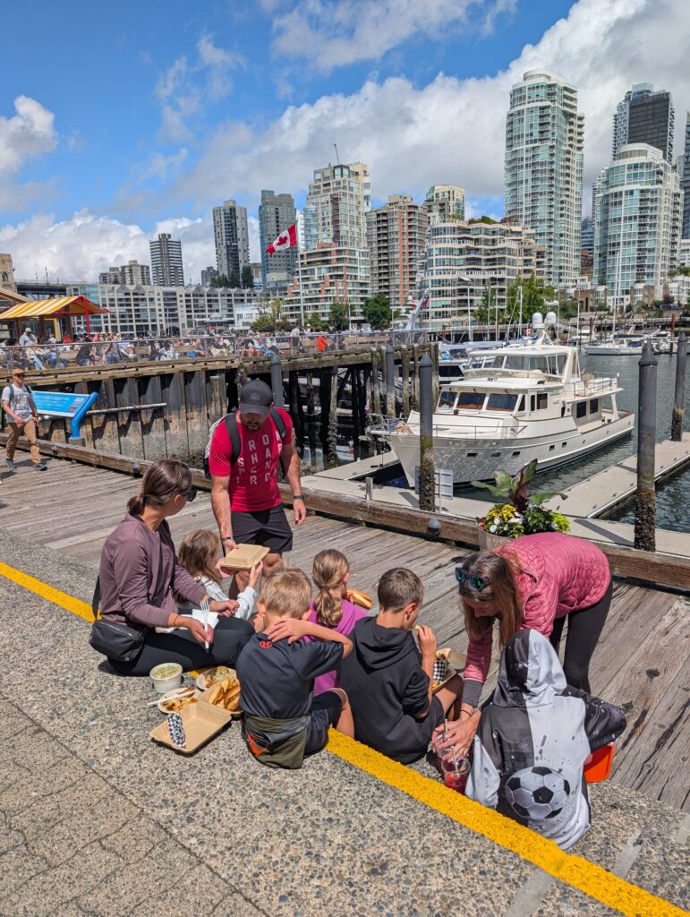 A group of people sitting on steps with boats, water, and skyscrapers behind them