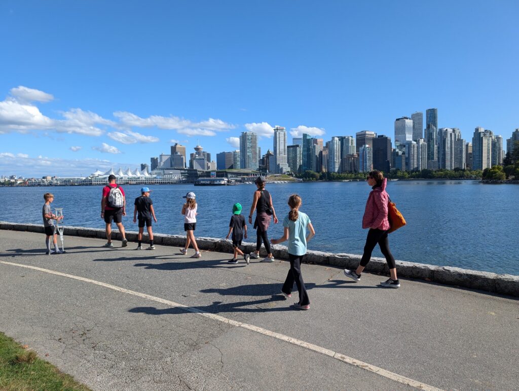 A group of people walking along a body of water on a paved path with the Vancouver skyline in the distance