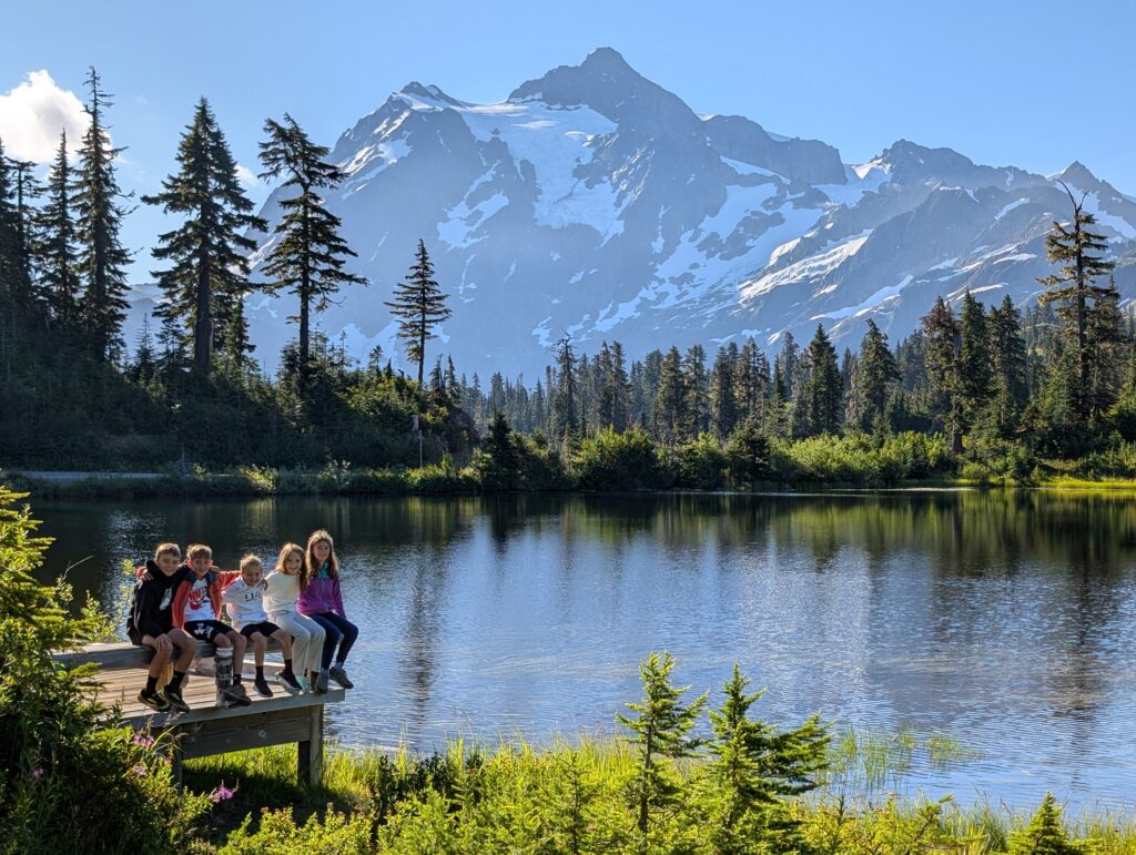 Kids sitting on a wooden bench near the edge of a pond with a big mountain in the background near Mt. Baker