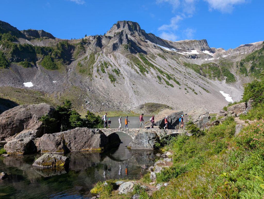 A group walking over a stone bridge with a rugged, rocky mountain peak behind them