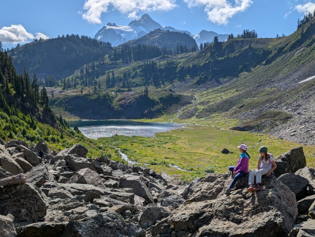 Two girls sitting on a large rock with a lake and tree-lined hills on the Chain Lakes Loop trail