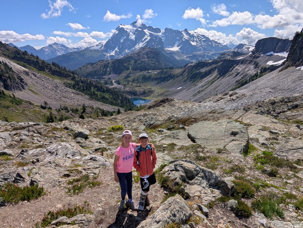 A young girl and boy standing on a rocky area with a lake, valley, and mountain behind them