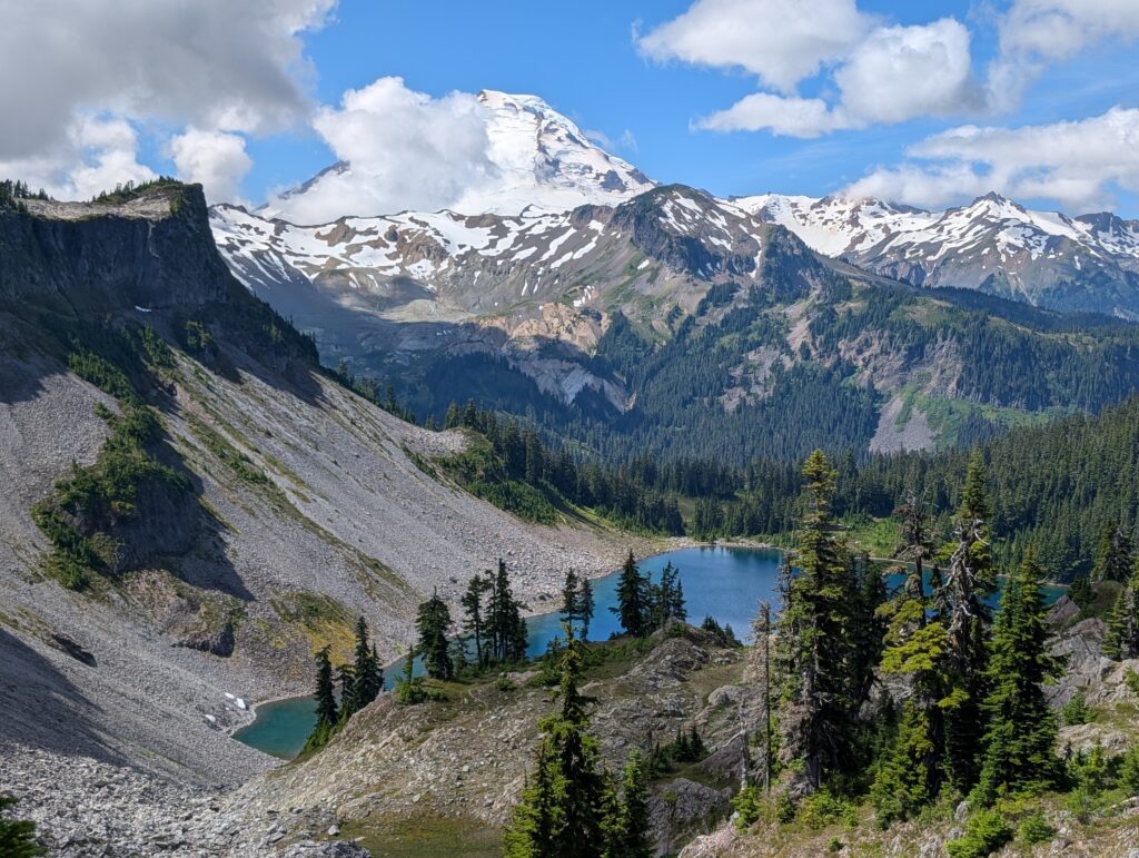 A prominent snowy mountain peak partially covered by clouds above a lake and trees