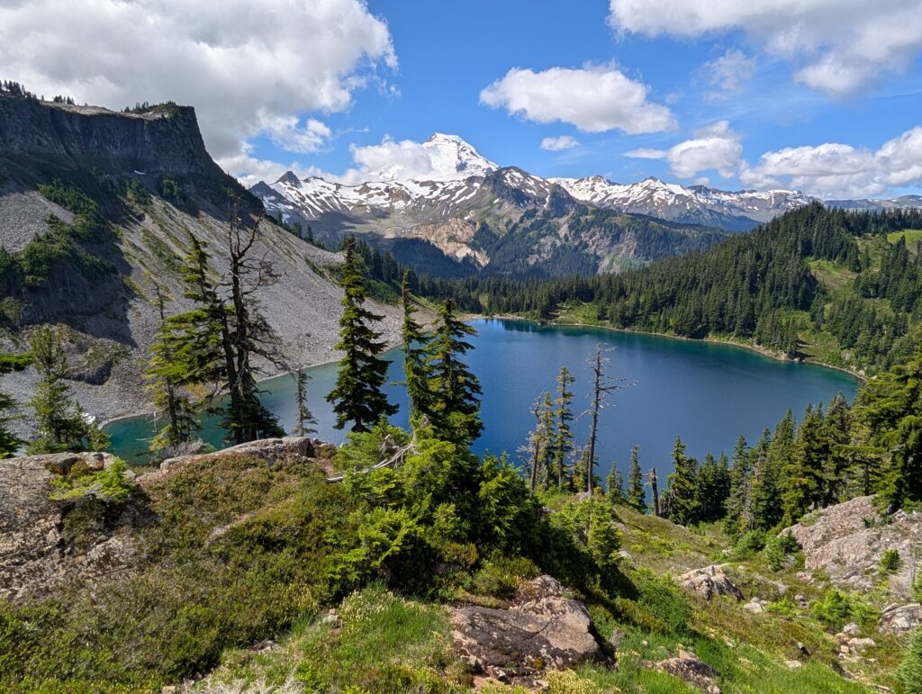 A larger blue-green lake with a green mound and trees in the foreground and snow-capped mountains behind it
