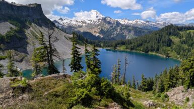 A larger blue-green lake with a green mound and trees in the foreground and snow-capped mountains behind it