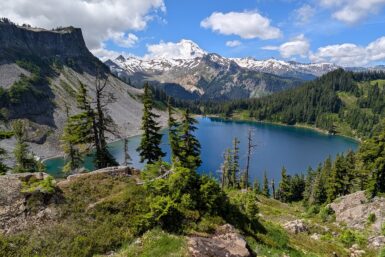 A larger blue-green lake with a green mound and trees in the foreground and snow-capped mountains behind it