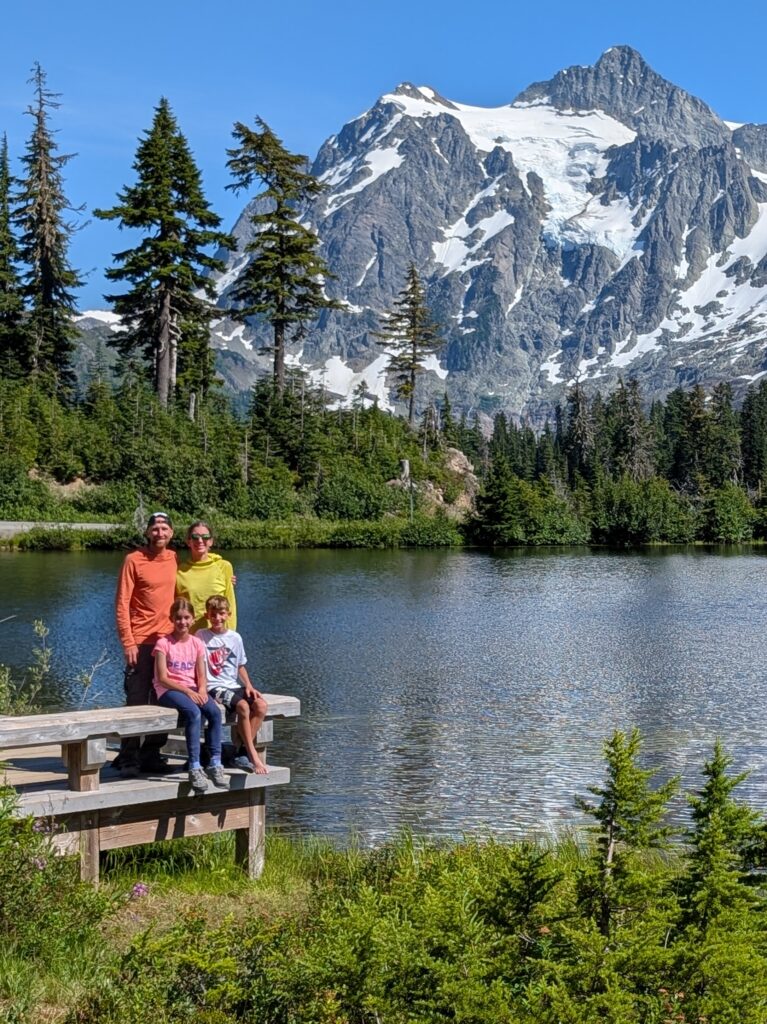 A family on a wooden platform next to a lake with a big mountain and trees behind them