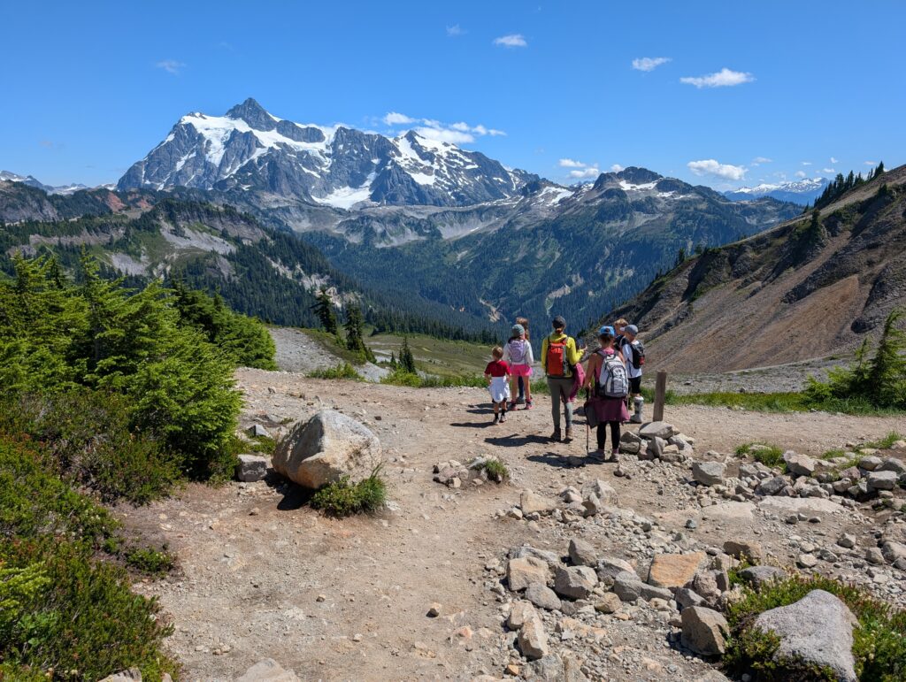 A group of hikers standing on a wide dirt path with a large mountain formation dotted with snow